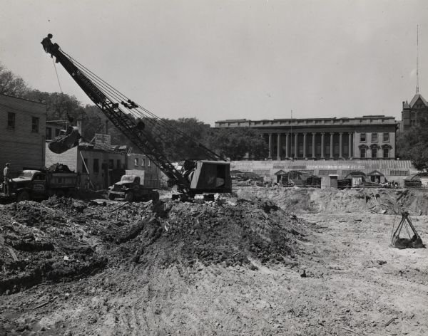 Library under construction on the University of Wisconsin-Madison campus. In the background is the State Historical Society, and Science Hall is in the upper right. Temporary metal-roofed quonset huts are on the site, with graffiti written on its side. A crane is just left of center. Gust K. Newburg Construction Co. is the name of the construction crew (on the left truck). The rear entrance for the Campus Grille is behind the vehicles.