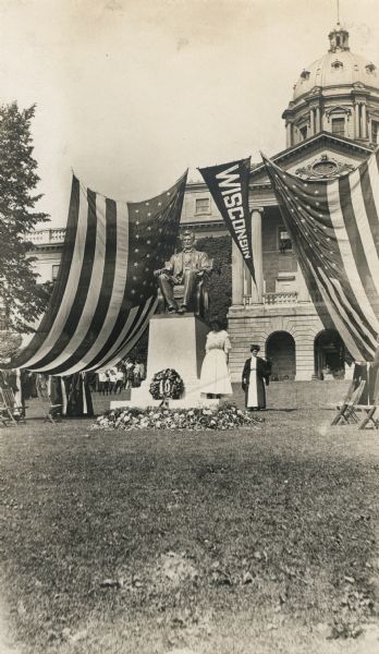 Unveiling ceremony, in front of Bascom Hall on the University of Wisconsin-Madison campus. Two American flags and a Wisconsin pennant hang from a wire above and behind the statue, depicting Lincoln sitting in a chair, on top of a plinth. Two women stand nearby, with a crowd in the background.