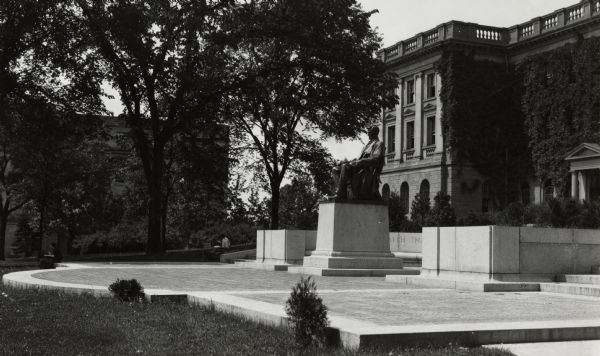 Side view of Lincoln Monument in front of Bascom Hall on the University of Wisconsin-Madison campus. A stone bench and paved walkway curves around the statue. Two men sit on the grass near the stairs on the left, and a woman sits on the edge of the paved area to the far left. In the background is Birge Hall.