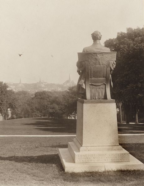 Rear view of Lincoln Monument on Bascom Hill on the University of Wisconsin-Madison campus. The back of the statue has a shield and eagle on the back of the chair, as well as other decorative details.  The base of the statue has state names carved into it. In the far distance is the isthmus with church steeples visible.