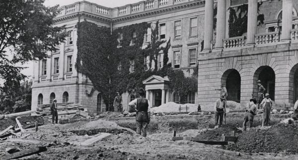 Lincoln Monument construction on the University of Wisconsin-Madison campus. Workmen are standing around the perimeter of a large hole while others are digging. A man near the front entrance of Bascom Hall is spraying water over the freshly dug dirt.