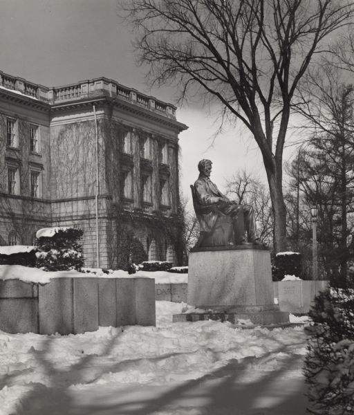 Side view of the Lincoln Monument on the University of Wisconsin-Madison campus. Bascom Hall is in the left background, and snow is on the ground. A stone bench and paved walkway curves around the statue, and the base of the statue has state names carved into it.