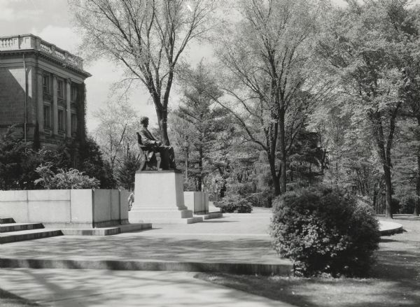 Side view of Lincoln Monument in front of Bascom Hall on the University of Wisconsin-Madison campus. A stone bench curves around the statue, which sits in a paved walk area. The base of the statue has state names carved into it