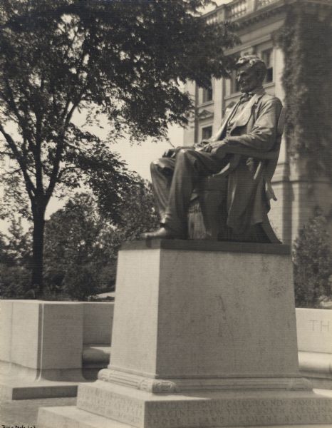Side view of Lincoln Monument on the University of Wisconsin-Madison campus. Bascom Hall is in the background. A stone bench curves around the statue, which sits in a paved walk area. The base of the statue has state names carved into it.