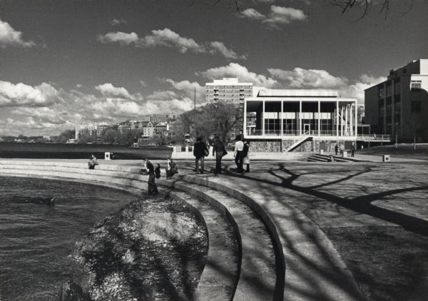 People are sitting or walking on the steps which curve along the shoreline at Memorial Union Terrace on the University of Wisconsin-Madison campus. The Alumni House is in the background, and in the far distance buildings line the Lake Mendota shoreline which curves around to the left.