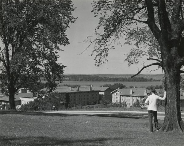View of dormitories on the University of Wisconsin-Madison campus. A man in a light-colored sweater is leaning with his arm against a tree on the right, looking towards buildings along the shoreline of Lake Mendota. In the far distance is Picnic Point.