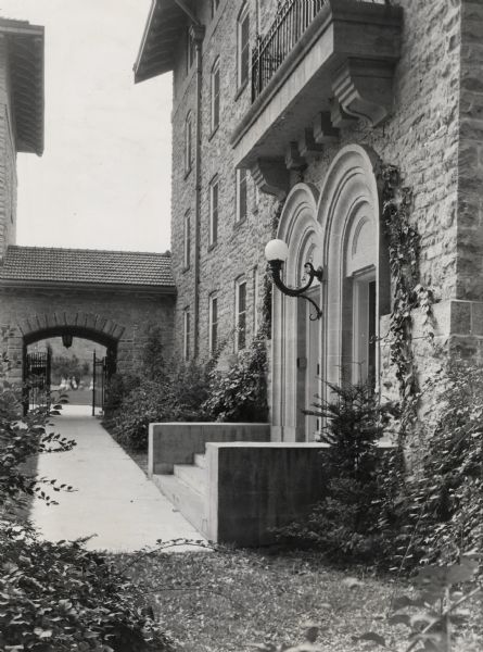 Entrance to a dormitory at the University of Wisconsin-Madison men's Residence Halls. A tiled-roofed arched gateway with wrought iron gates linking the dormitory to the Tripp quadrangle and another building is in the background. A small group of people can be seen sitting in a courtyard area in the far background.