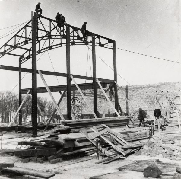 Men working on construction of dormitories on the University of Wisconsin-Madison campus. Construction works are on top of a partially constructed frame and working on the ground amongst piles of materials.  A stone wall with a framed-up arch is in the background. In the far background are trees along the shoreline of Lake Mendota.