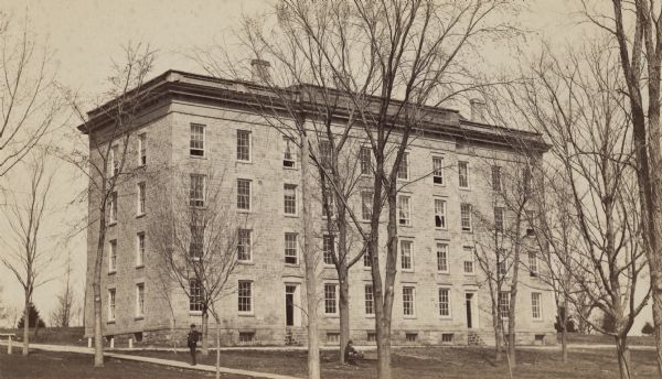 View up Bascom Hill of North Hall dormitory on the University of Wisconsin-Madison campus. Two people are outside, one standing on the sidewalk, the other sitting on a bench near a tree.