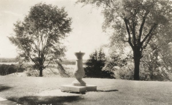 A sundial on a pedestal sits on Observatory Hill, overlooking trees, a rooftop, and part of Lake Mendota.