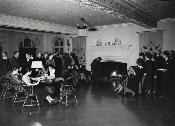 Interior room of student center on the University of Wisconsin-Madison campus. A student meeting is being held. One man stokes the fire in the fireplace, other people sit in couches flanking the fireplace, while others mill around the edges of the room. A group of women sit at a table on the left, talking and reading articles.