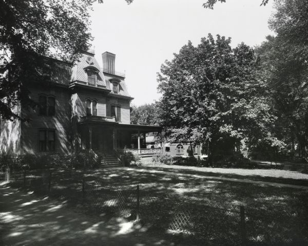Residence on the University of Wisconsin campus. Later this site became the Memorial Union. A fence and a drive run along the bottom of the image toward the left. Another building is to the right of the house, partially hidden behind the trees.