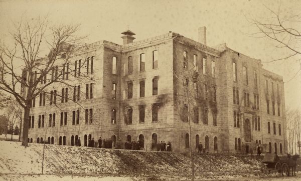 View across Park Street of Science Hall building after the fire of December 1, 1884, on the University of Wisconsin-Madison campus. The building's exterior is discolored from the smoke, and snow is on the ground. People in heavy coats and hats are gathered along the left side and front of the building. A horse-drawn buggy stands in the lower right corner.