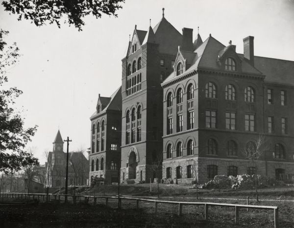 Exterior of Science Hall on the University of Wisconsin-Madison campus. View from across Park Street taken shortly after completion of new building after fire of Old Science Hall in 1884. Music Hall is in the background on the other side of Bascom Hill. A wood fence is along Park Street which is dirt, and building supplies are stacked around the foundation of Science Hall.