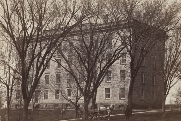 View across Bascom Hill of South Hall on the University of Wisconsin-Madison campus. There is a horse-drawn vehicle under a tree, and a man is working on the lawn. Another man walks near the horse.