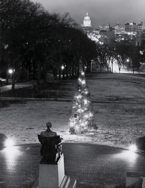 Elevated view looking east from Lincoln Terrace on Bascom Hill on the University of Wisconsin-Madison campus. The Lincoln Monument is in the foreground and a lighted Christmas tree is on the snow-covered lawn in front of it. In the distance is State Street leading to the Wisconsin State Capitol.