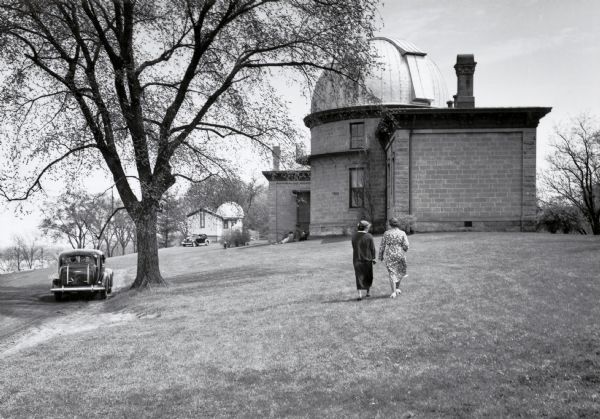 Two women are walking on the lawn towards the observatory building on the University of Wisconsin-Madison campus. A car is parked on the lawn near a tree on the left, and part of Lake Mendota is on the far left.