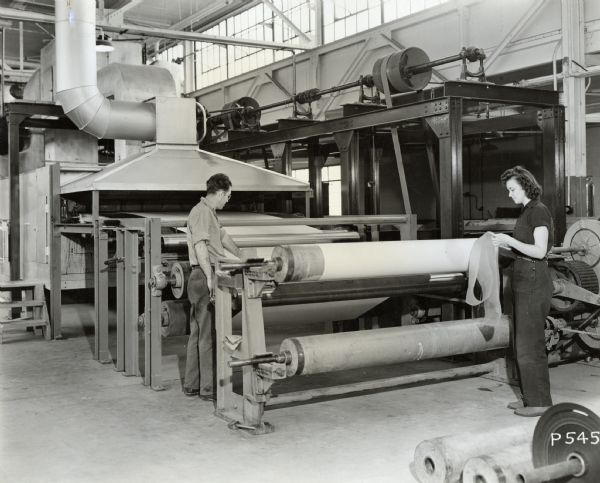 Two employees of the Consolidated Paper Company check the quality of the laminate product emerging from the dry end of the treater.  The laminate was manufactured as part of the company's war work for the military. The man in the photograph has been identified as Bob Schutz; the woman is unidentified.