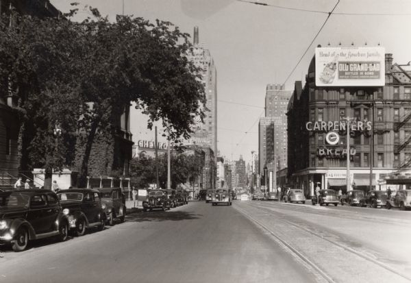Looking east on Wisconsin Avenue in the 800 block between 8th and 9th Streets. Cars are parked and driving down the street. An apartment block called the Belvedere was located on the southeast corner of 8th and Wisconsin. The Milwaukee Public Library is partially hidden behind trees in the left foreground. Signs advertise Carpenter's Bread, Old Grand-Dad (liquor), and Chevrolet (automobile).