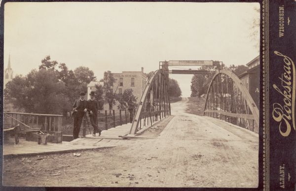 Two men standing on a dirt road at a bridge with the Nichols House in the background.
