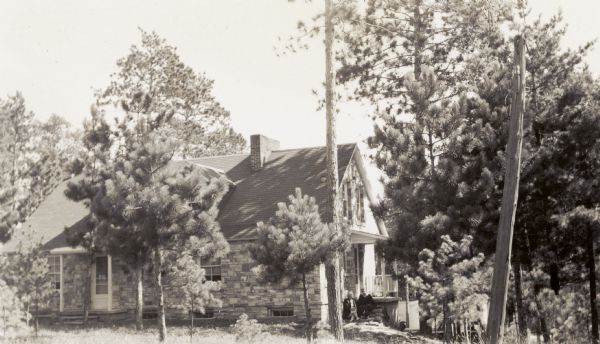 Exterior view of the stone-walled headquarters building of the Water Regulatory Board located within a grove of trees. According to text included with the photograph, the main building is 42' wide and 27' long and includes a basement garage and work room, well room, office and furnace room. Above the garage room and work shop is a six room modern apartment for the use of an employee of the department, and a three room field office and apartment. A man and woman sit on the steps.