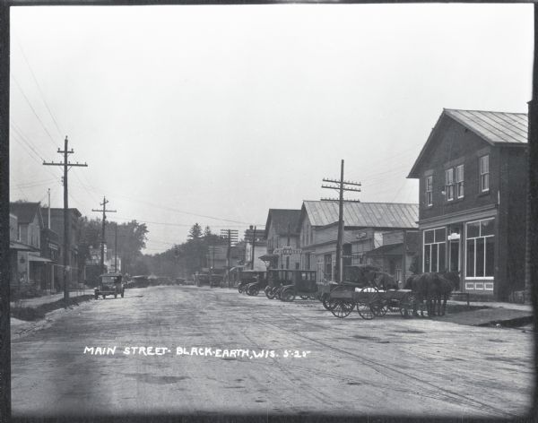 Main Street with storefronts, automobiles, and horses and carriages parked along it.