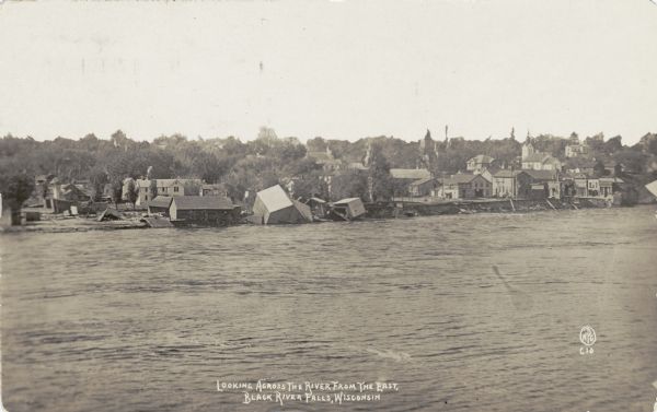 View looking across the river east into Black River Falls during the flood, showing the elevated level of the river and buildings falling into the water. Caption reads: "Looking Across the River from the east, Black River Falls, Wis."
