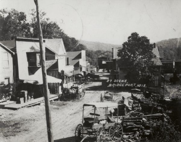 Elevated view of street, with horse-drawn vehicles parked in front of storefronts. A covered bridge is in the background.