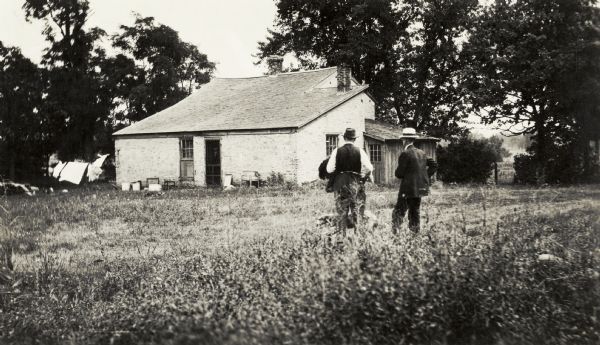 Exterior view of the Strang Cottage, with two men and a dog standing in front of it. Laundry hangs on a line behind the cottage.