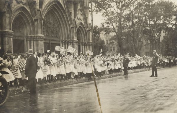 A group of women and girls wearing white dresses and waving American flags are standing at the curb in front of a church. Three police officers are standing in the street.