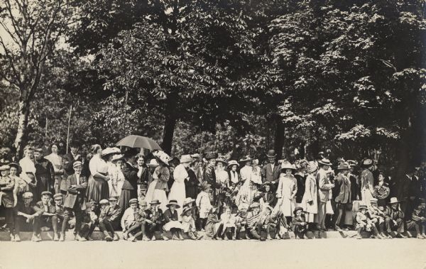 A crowd of spectators standing and sitting on a curb waiting for a parade.