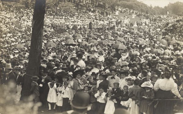 Elevated view of a large crowd of women, children, and men filling a field.  A rope strung between trees creates a barrier in the foreground.