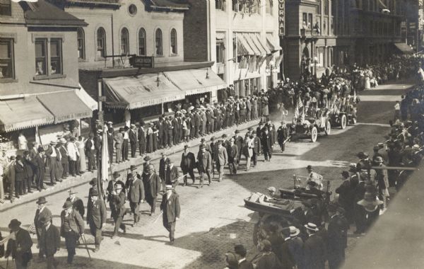 Elevated view of a group of men marching down the center of the road with two cars decorated with flags following close behind. Both sidewalks are full of people.