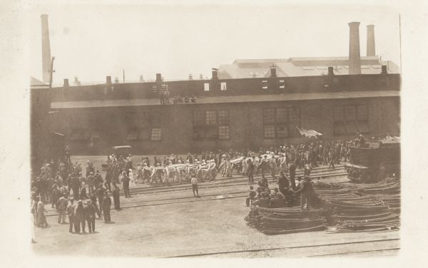 Elevated view of a parade crossing through a factory yard, where the workers are watching. Some are sitting on the roof of the building in the background. The parade participants are carrying two large flags, with three smaller ones on poles behind.