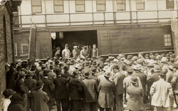 A crowd has gathered around an open door on a boat from which several men are reaching out and shaking hands with individuals in the crowd.