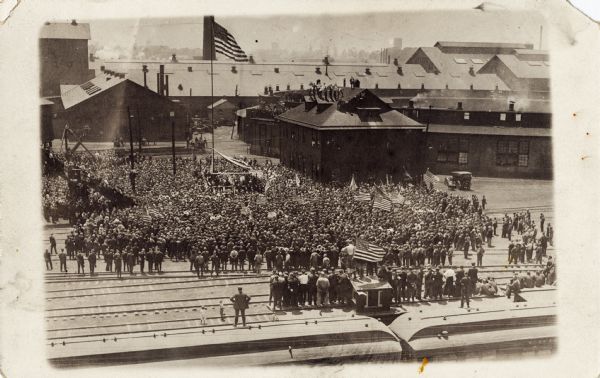 Elevated view of large crowd gathered in a railroad yard. Men are gathered around a podium in the middle of the crowd.