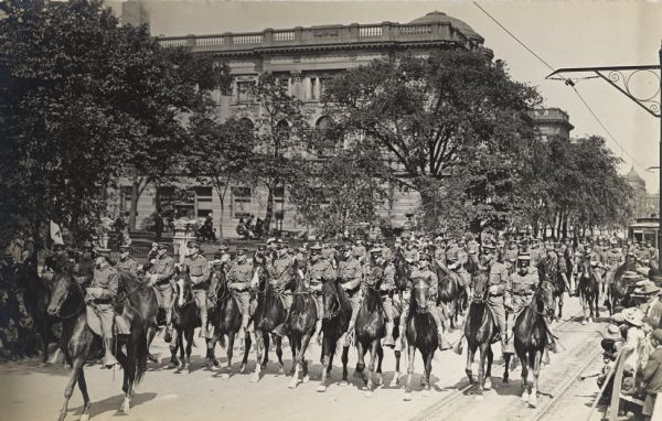 A group of soldiers on horseback in a parade. The road is lined with people.