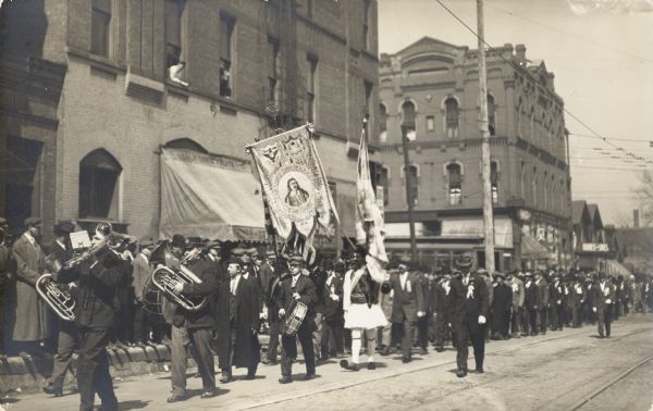 A small band leads a group of men with ribbons on their jackets.  In between are two men in folk costumes carrying two banners.  A crowd watches from the sidewalk.
