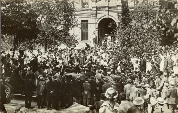 Theodore Roosevelt raising his hat amidst a crowd in Milwaukee. He is leaving a building and is heading toward a waiting car.
