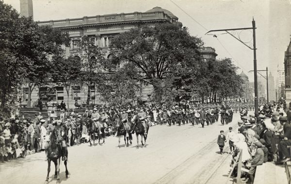 Street parade led by four men on horses with a marching band following. A crowd is gathered to watch the parade. People are sitting on benches in a park on the left near a large building.
