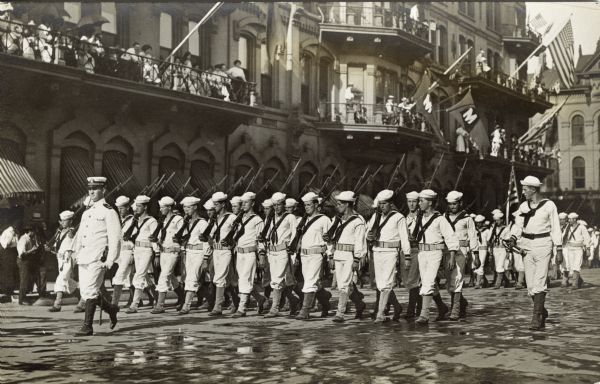 A group of sailors marching with rifles in a parade while onlookers are watching from the balconies of a large building in the background.