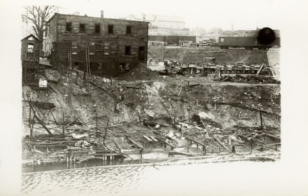 Elevated view across water towards a building at the top of a steep incline, with a small group of people to the left. On top of the hill to the right are railroad cars on tracks. There is debris along the steep shoreline.