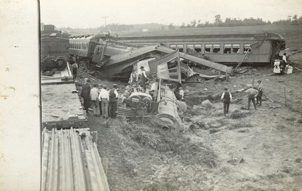 Elevated view of a Northwestern Line railroad accident. Railroad cars have fallen off the railroad tracks and the engine car is lying on its side. Men and women are standing around and on top of the locomotive.