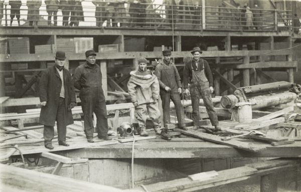 Three men on pilings posing with man in a diving suit in preparation for a dive. Onlookers are standing above on a bridge in the background.