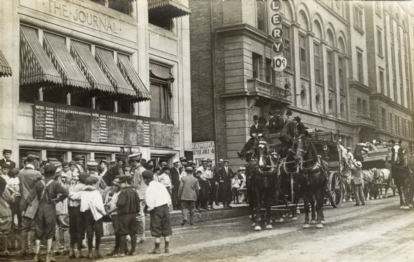 Heavy touring carriages unload passengers in front of the <I>Milwaukee Journal</I> newspaper building. A crowd is gathered, along with a few musicians and paperboys. Over the entrance to the building is a baseball scoreboard listing scores for each inning of a baseball game, as well as the battery (pitcher and catcher) for each team.