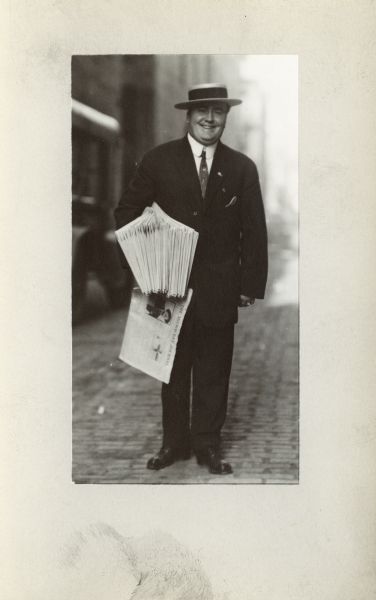 Man in a suit and hat standing on the street holding a stack of <i>Milwaukee Journal</i> newspapers.