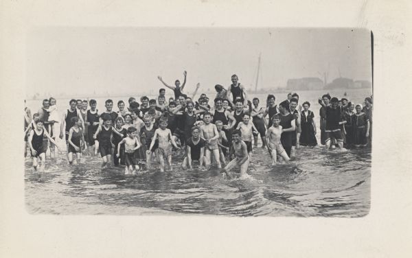 A large group of men, women, and children, wearing swimming attire, standing in a lake. Industrial buildings are in the distance along the shoreline.