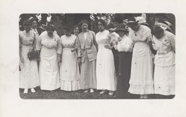 A row of eight woman wearing white dresses and standing outdoors. They appear to be playing a game involving holding a string in their mouth with a weight hanging from the end.