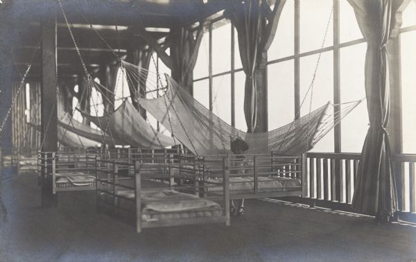 A little girl is sitting on a hammock on an enclosed porch with large windows. There are also several wooden and padded hanging seats.
