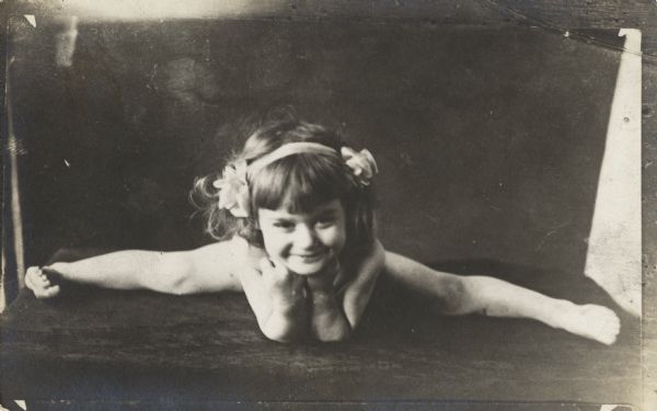 A little girl is on the floor doing the splits resting her head on her hands. She has a ribbon with flowers in her hair.